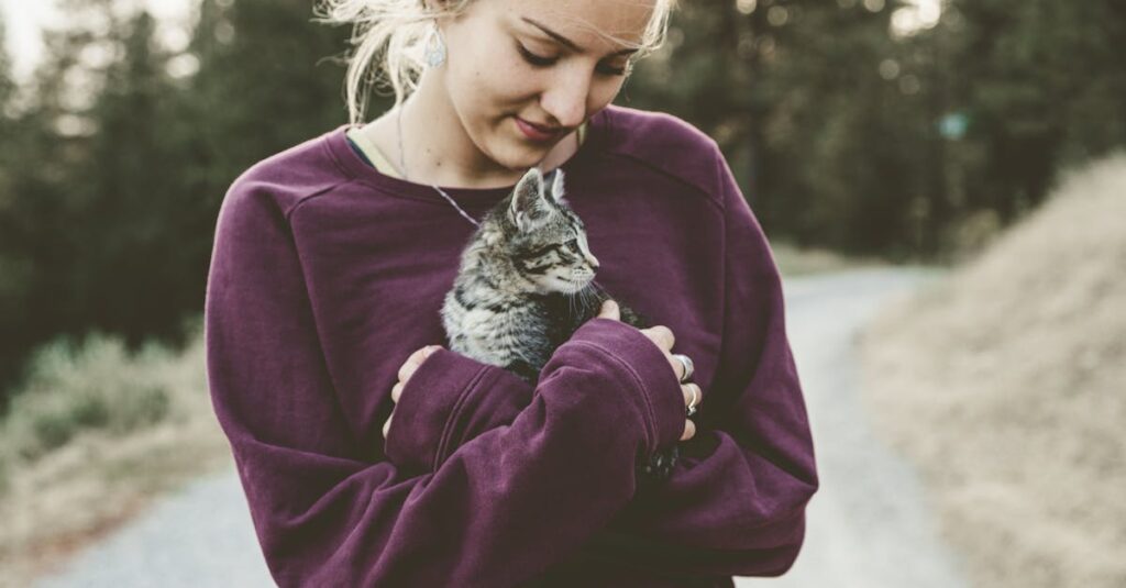 Young woman in a purple sweater smiling down at a kitten she's holding outdoors on a pathway.