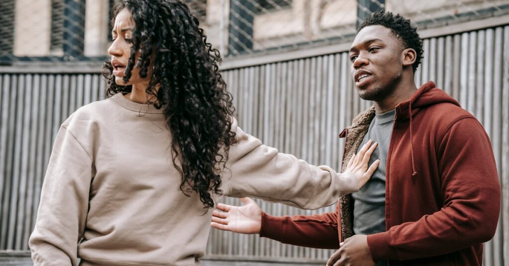 A couple in casual clothing appears to be in a disagreement outdoors with grey fencing background.