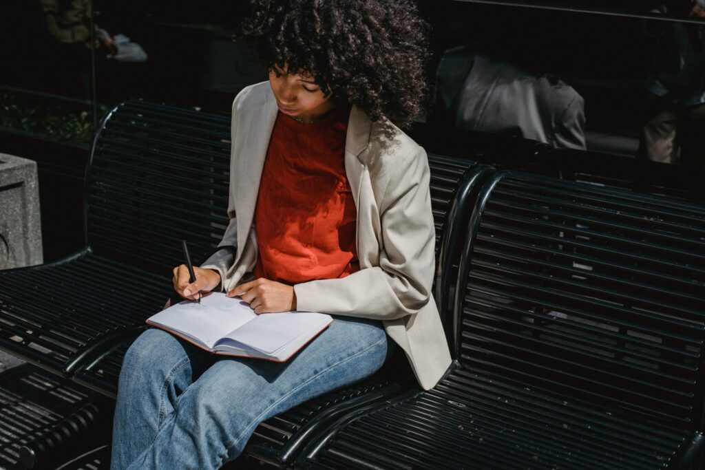 A young woman sits on a bench outdoors writing in a notebook during the day.