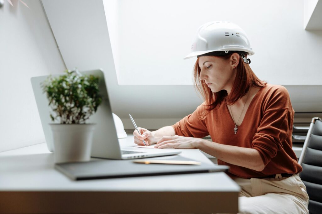 A focused female architect in a hard hat writing notes while working on a laptop indoors.