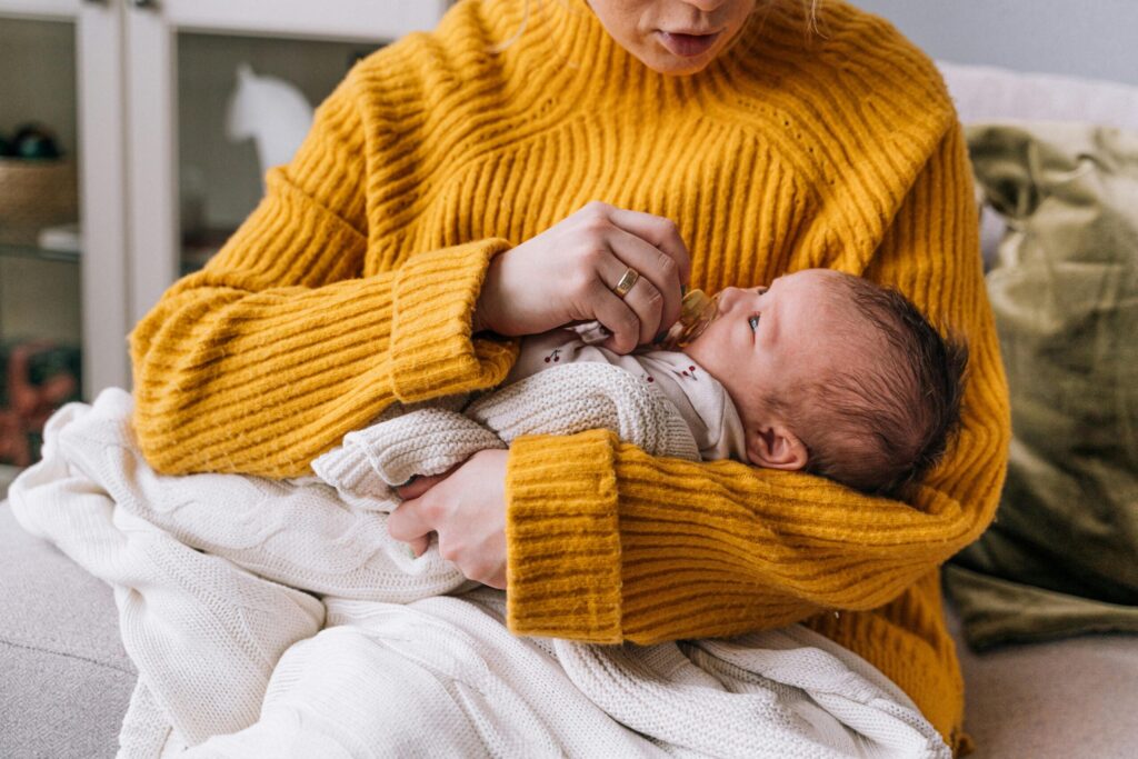 A mother lovingly holds her newborn baby, wrapped in a blanket, wearing a yellow sweater indoors.