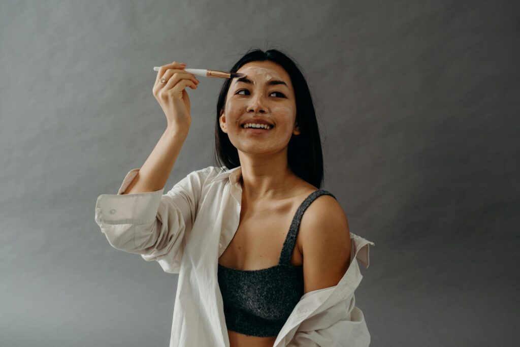 Smiling Asian woman applying facial cream with a brush, promoting beauty and skincare in a studio setting.