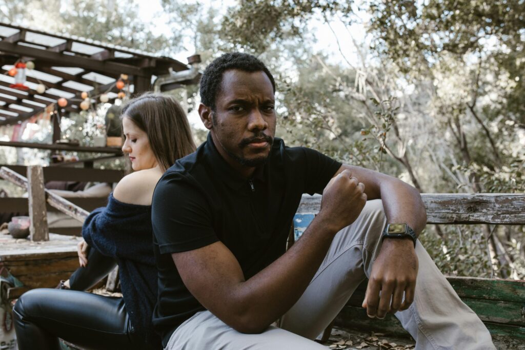 A thoughtful man sits outdoors, with a woman sitting distantly behind him.