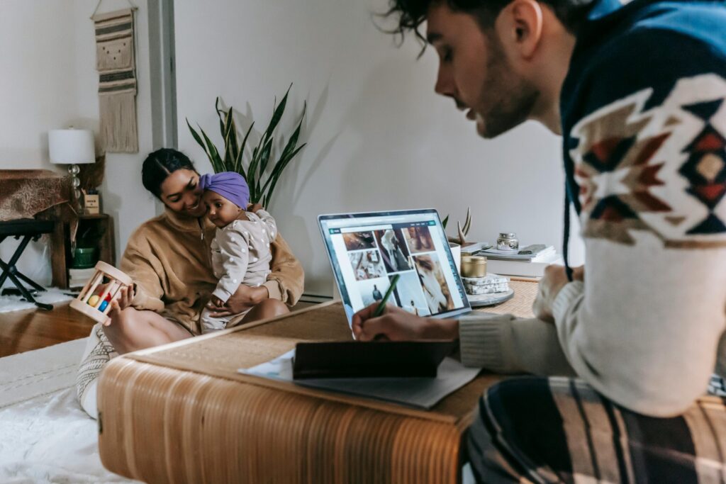 Crop pensive ethnic father writing in notebook and working with laptop while ethnic mother playing with cute little daughter