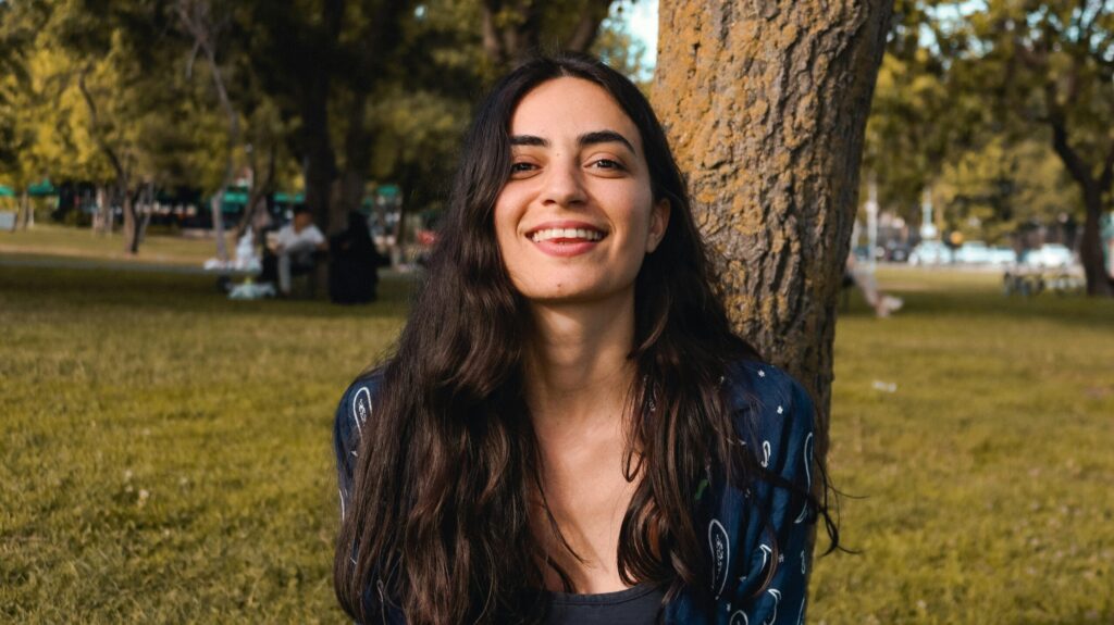 Smiling young woman leaning against a tree in a sunlit park.