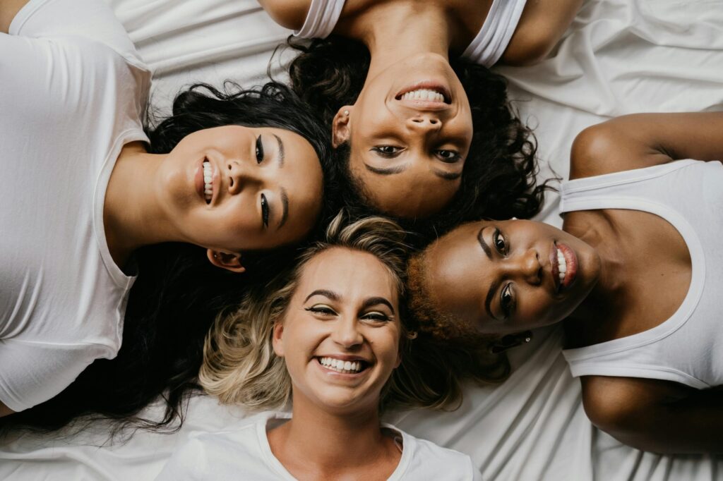 Four diverse women lying on a bed smiling at the camera, sharing a joyful moment.