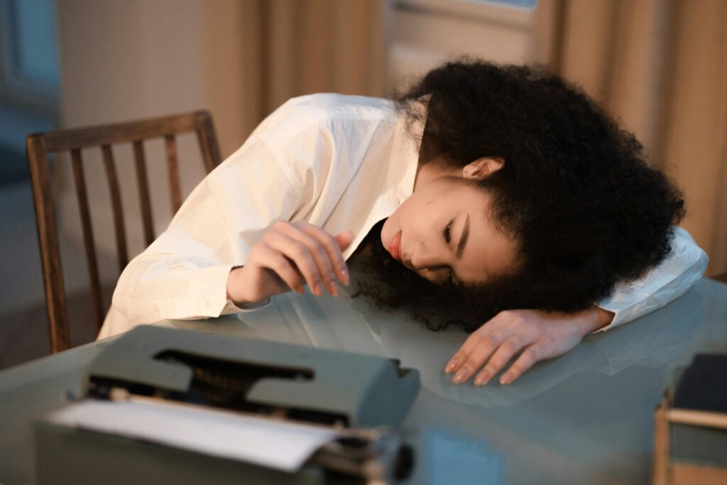 A woman with afro hair in a white shirt rests her head on a table next to a vintage typewriter, appearing fatigued.