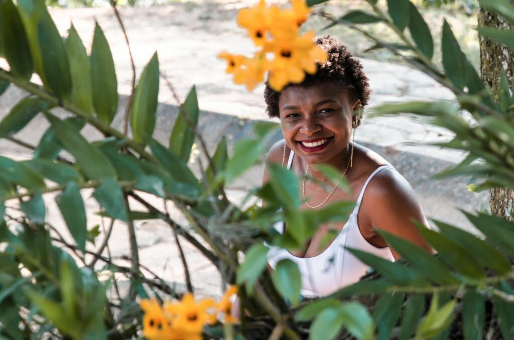 Joyful woman smiling behind vibrant yellow flowers surrounded by greenery.