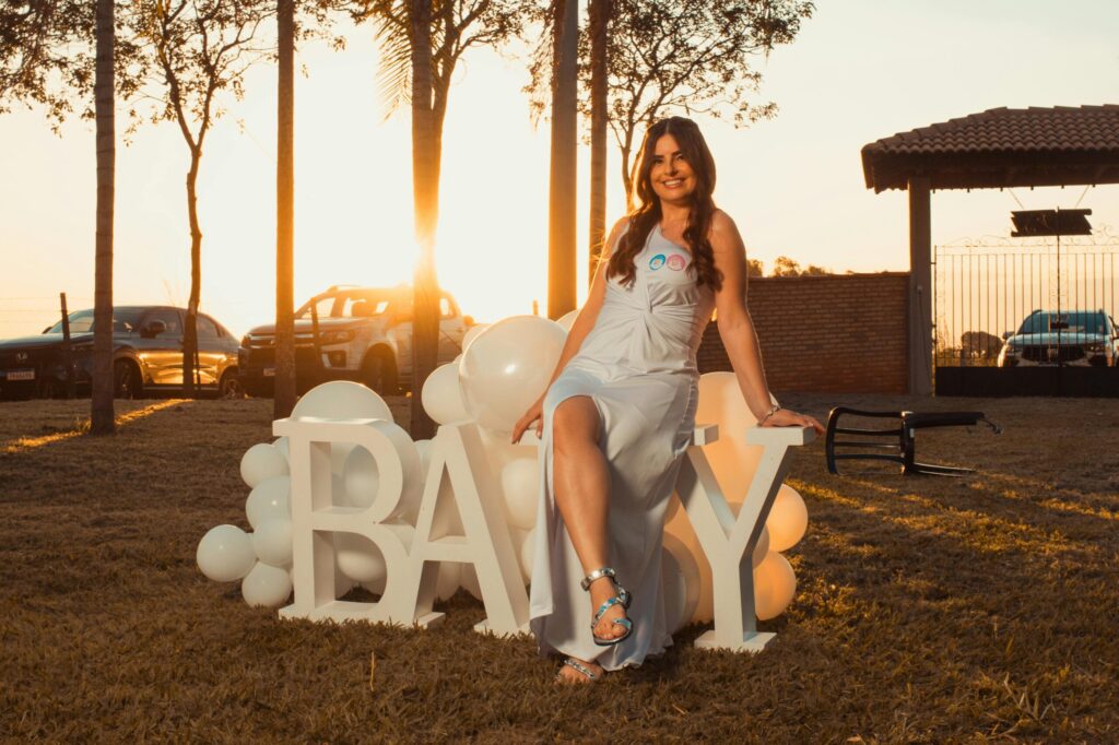 A woman sitting on large baby letters with balloons during sunset. Outdoor celebration with a warm glow.