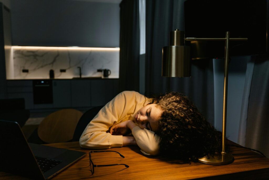 A tired woman with curly hair asleep at a desk with a laptop and glasses under warm lamp light in a dimly lit room.