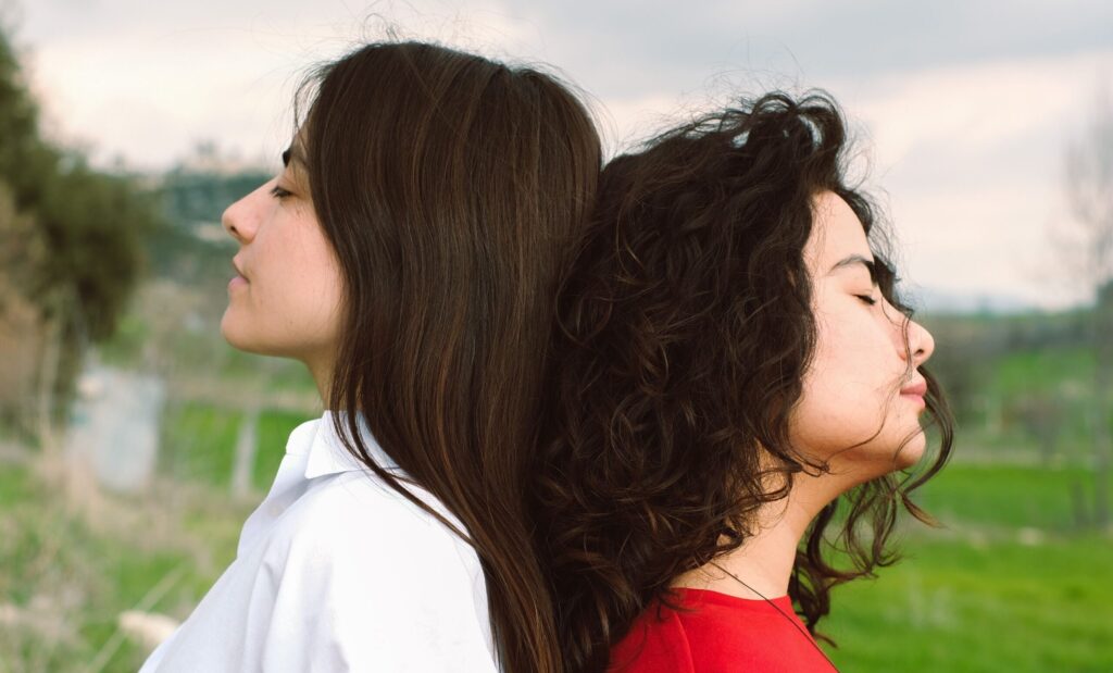 Two women stand back-to-back, eyes closed, in serene countryside setting.