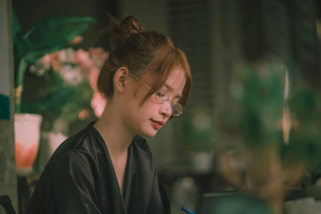 Young woman with glasses relaxing indoors, surrounded by plants and warm light.